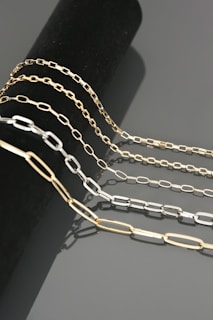 Four chains of varying lengths and colors drape elegantly over a cylindrical black surface. The chains have a sleek, metallic finish and are composed of rectangular links. The reflections on the smooth gray background add to the luxurious appearance.