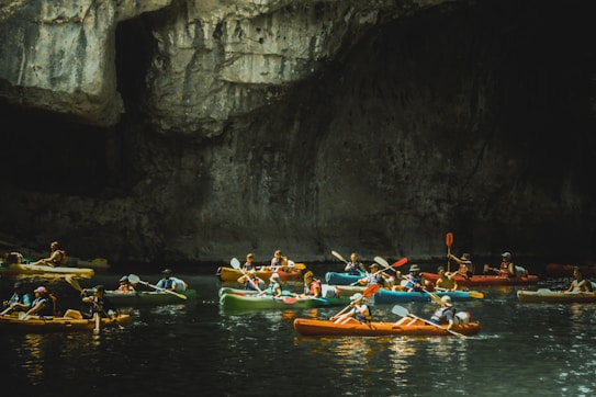 A group of people are kayaking inside a dimly lit cave. The scene is lively with several colorful kayaks, and the dark, textured stone walls of the cave contrast with the water below.