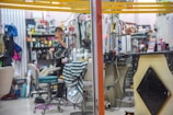 A cluttered interior of a beauty salon or barber shop with various grooming products on shelves. A person is eating an apple while standing near the counter, and various salon instruments and chairs are visible. A small cat is partially visible at the bottom right corner.