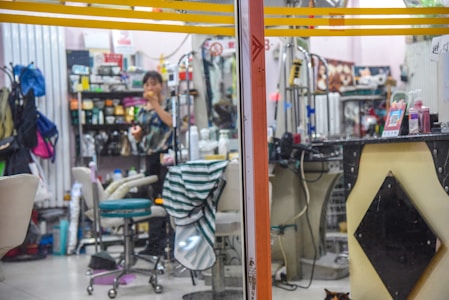 A cluttered interior of a beauty salon or barber shop with various grooming products on shelves. A person is eating an apple while standing near the counter, and various salon instruments and chairs are visible. A small cat is partially visible at the bottom right corner.