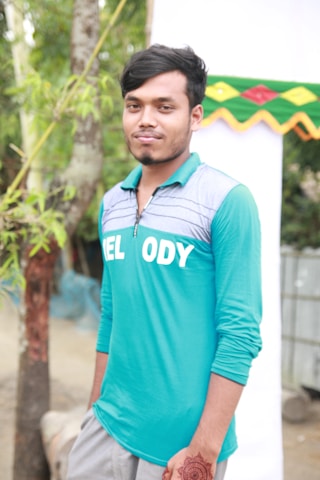 A young man with short dark hair stands outdoors, wearing a turquoise shirt with a collar and the word 'MELODY' written on it. He has a calm expression and resting against his side is a hand with henna designs. In the background, there's greenery and a tall tree, along with a partially visible white and green festive decoration.