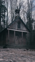 A small, rustic wooden chapel surrounded by dense, leafless trees. The structure features a simple design with a steep roof and a belfry. The wood is weathered, giving it an aged appearance. A stone pathway leads up to the chapel, adding to its quaint charm.