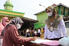 Two women, both wearing headscarves and face masks, are engaged in a discussion at an outdoor registration table. One woman is standing and appears to be overseeing, while the other is seated and writing on a document. In the background, several people are queued up near a green and yellow building with a domed roof, likely a mosque. A banner advertising free medical treatment is visible.