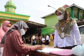 Two women, both wearing headscarves and face masks, are engaged in a discussion at an outdoor registration table. One woman is standing and appears to be overseeing, while the other is seated and writing on a document. In the background, several people are queued up near a green and yellow building with a domed roof, likely a mosque. A banner advertising free medical treatment is visible.