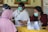 Several people wearing face masks are seated around a table, engaged in an interaction or discussion. One person appears to be filling out forms while another uses a smartphone. On the table, there are various hygiene products such as hand sanitizer and tissue boxes, indicating a setting mindful of health precautions.