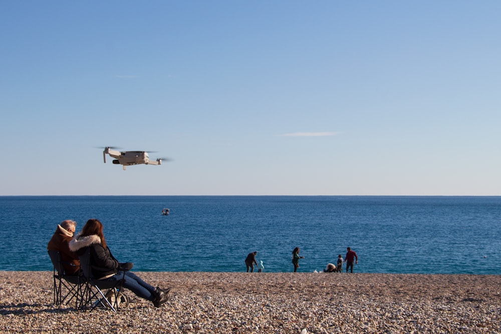 a couple of people sit on a beach watching a plane fly by