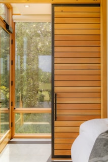 A modern interior design featuring a large wooden sliding door with horizontal planks and a black frame. Large windows allow natural light to stream in, with lush greenery visible outside. Part of a cozy bed with white sheets and a gray blanket is visible in the foreground.