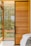 A modern interior design featuring a large wooden sliding door with horizontal planks and a black frame. Large windows allow natural light to stream in, with lush greenery visible outside. Part of a cozy bed with white sheets and a gray blanket is visible in the foreground.