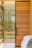 A modern interior design featuring a large wooden sliding door with horizontal planks and a black frame. Large windows allow natural light to stream in, with lush greenery visible outside. Part of a cozy bed with white sheets and a gray blanket is visible in the foreground.