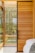 A modern interior design featuring a large wooden sliding door with horizontal planks and a black frame. Large windows allow natural light to stream in, with lush greenery visible outside. Part of a cozy bed with white sheets and a gray blanket is visible in the foreground.