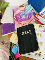 A cluttered tabletop features a variety of colorful notebooks and journals. One prominent black notebook has the word 'IDEAS' on it. Other visible items include a colorful journal with the phrase 'don't overthink it' and a remote control. Brightly colored pens are scattered among the notebooks.