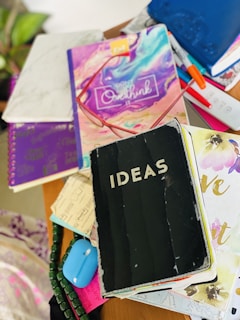 A cluttered tabletop features a variety of colorful notebooks and journals. One prominent black notebook has the word 'IDEAS' on it. Other visible items include a colorful journal with the phrase 'don't overthink it' and a remote control. Brightly colored pens are scattered among the notebooks.
