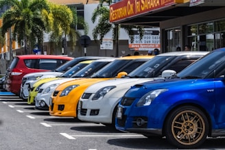 A row of colorful cars parked beside each other in a parking lot, with palm trees and a building featuring a restaurant sign in the background.