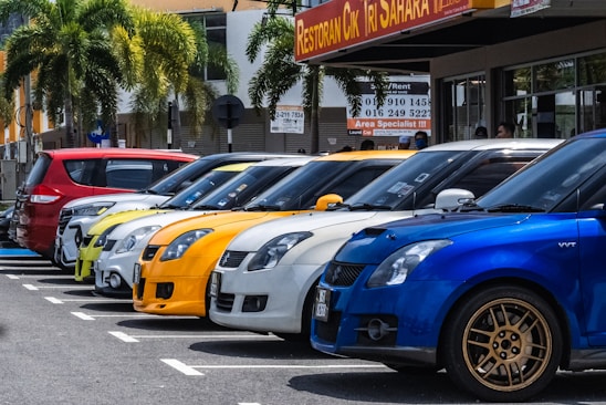 A row of colorful cars parked beside each other in a parking lot, with palm trees and a building featuring a restaurant sign in the background.