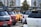 A parking lot filled with several Suzuki cars in various colors including red, yellow, and white. The vehicles have spoilers and show urban buildings and trees in the background under a partly cloudy sky.