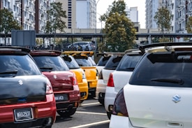 A parking lot filled with several Suzuki cars in various colors including red, yellow, and white. The vehicles have spoilers and show urban buildings and trees in the background under a partly cloudy sky.