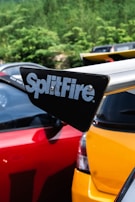 A close-up view of a car's rear spoiler featuring the brand name 'SplitFire.' The spoiler is mounted on a yellow car, with a red car parked next to it. The background consists of green foliage and parts of other vehicles are visible.