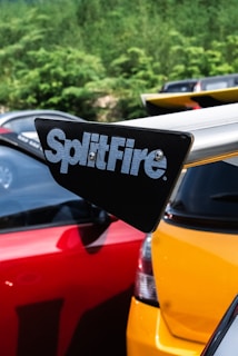 A close-up view of a car's rear spoiler featuring the brand name 'SplitFire.' The spoiler is mounted on a yellow car, with a red car parked next to it. The background consists of green foliage and parts of other vehicles are visible.