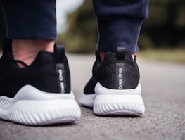A close-up view of the back of someone's feet wearing black and white athletic shoes. The shoes have a sleek design with textured white soles and a black mesh fabric. The person is standing on an outdoor surface, possibly pavement.