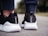 A close-up view of the back of someone's feet wearing black and white athletic shoes. The shoes have a sleek design with textured white soles and a black mesh fabric. The person is standing on an outdoor surface, possibly pavement.