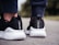 A close-up view of the back of someone's feet wearing black and white athletic shoes. The shoes have a sleek design with textured white soles and a black mesh fabric. The person is standing on an outdoor surface, possibly pavement.
