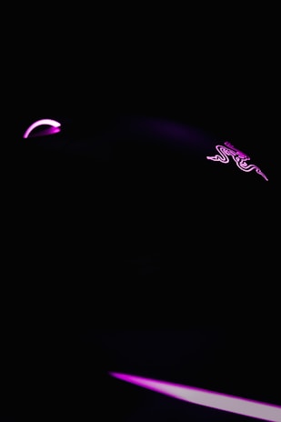 A dark, minimalist scene featuring a gaming mouse highlighted with neon pink lighting. The mouse logo is faintly visible, and the overall ambiance is sleek and futuristic.