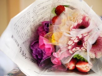 A colorful bouquet of flowers wrapped in a newspaper. The vibrant mix includes pink and purple blossoms, with touches of red, yellow, and orange. Delicate petals and green leaves are visible through the translucent wrapping.