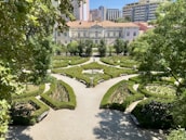 An intricately designed garden with symmetrical hedges forming geometric patterns, surrounded by trees and with a path leading to a large, elegant building in the background. Tall apartment buildings loom over the scene, adding an urban contrast to the landscaped garden.