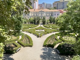 An intricately designed garden with symmetrical hedges forming geometric patterns, surrounded by trees and with a path leading to a large, elegant building in the background. Tall apartment buildings loom over the scene, adding an urban contrast to the landscaped garden.