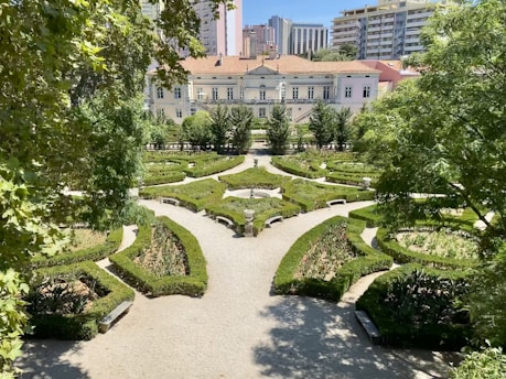 An intricately designed garden with symmetrical hedges forming geometric patterns, surrounded by trees and with a path leading to a large, elegant building in the background. Tall apartment buildings loom over the scene, adding an urban contrast to the landscaped garden.