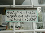 A humorous sign with playful text hangs on a glass window, surrounded by other decorative items. The sign reads, 'At the beach we don't hide crazy... We parade it out on the porch and hand it a cocktail!' The window frames are white, and there are visible reflections from outside.