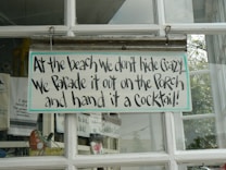 A humorous sign with playful text hangs on a glass window, surrounded by other decorative items. The sign reads, 'At the beach we don't hide crazy... We parade it out on the porch and hand it a cocktail!' The window frames are white, and there are visible reflections from outside.