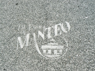 White text and a symbol are stenciled on a gray, speckled asphalt surface. The words appear to include 'The Town of Manteo' along with a circular emblem featuring a building-like figure.