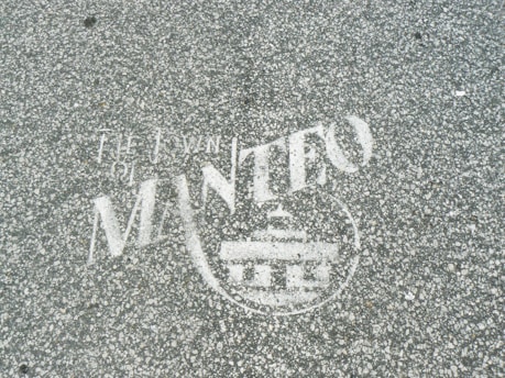 White text and a symbol are stenciled on a gray, speckled asphalt surface. The words appear to include 'The Town of Manteo' along with a circular emblem featuring a building-like figure.