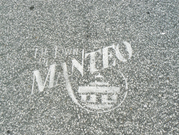 White text and a symbol are stenciled on a gray, speckled asphalt surface. The words appear to include 'The Town of Manteo' along with a circular emblem featuring a building-like figure.