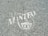 White text and a symbol are stenciled on a gray, speckled asphalt surface. The words appear to include 'The Town of Manteo' along with a circular emblem featuring a building-like figure.