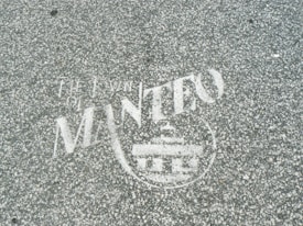 White text and a symbol are stenciled on a gray, speckled asphalt surface. The words appear to include 'The Town of Manteo' along with a circular emblem featuring a building-like figure.