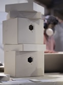 A stack of plain, cardboard boxes with hexagonal cutouts is on a table. In the background, a person wearing headphones and a mask is visible, likely engaging in a craft or work activity.