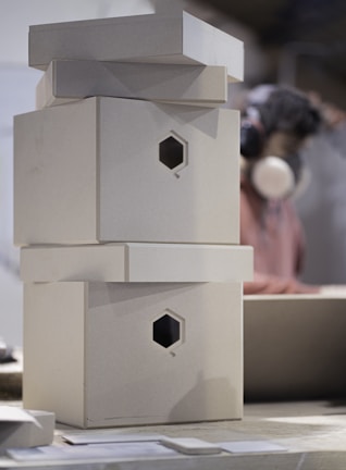 A stack of plain, cardboard boxes with hexagonal cutouts is on a table. In the background, a person wearing headphones and a mask is visible, likely engaging in a craft or work activity.