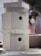 A stack of plain, cardboard boxes with hexagonal cutouts is on a table. In the background, a person wearing headphones and a mask is visible, likely engaging in a craft or work activity.