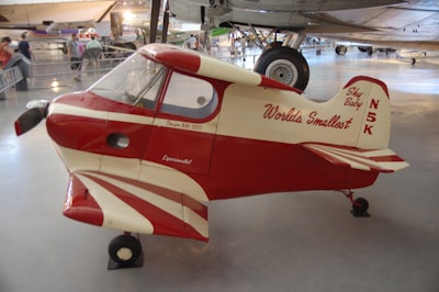 A small, red and white aircraft labeled as 'World's Smallest' with the tail number N5K, displayed in a museum environment with other planes and people in the background.