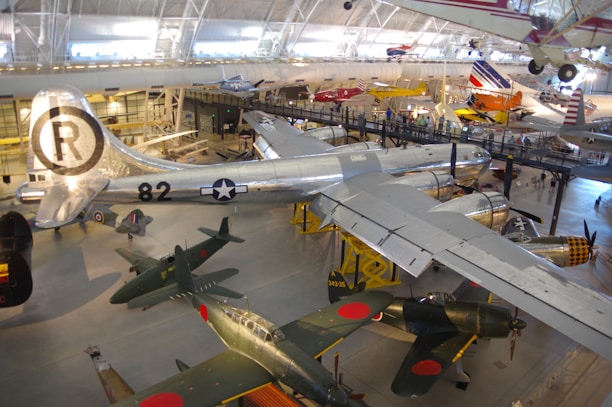An indoor aviation museum displays a variety of historic airplanes. The large metallic aircraft with the number 82 and a star insignia stands out in the center. Smaller planes with distinct markings and designs are situated around it. The museum environment is well-lit, with people observing the exhibits on an elevated walkway.