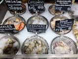 A variety of seafood displayed on ice in metal trays includes stone crab claws, littleneck clams, jumbo scallops, cockles, steamers, and squid. Each type of seafood is accompanied by a blackboard sign indicating its origin and price.