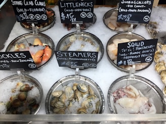 A variety of seafood displayed on ice in metal trays includes stone crab claws, littleneck clams, jumbo scallops, cockles, steamers, and squid. Each type of seafood is accompanied by a blackboard sign indicating its origin and price.