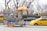 A street-side halal food cart offering a variety of items like hot dogs and falafel. The cart is adorned with colorful food images and a bright umbrella. It is parked on a city street next to a yellow taxi and a gray car, with leafless trees and a stone wall in the background.