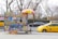 A street-side halal food cart offering a variety of items like hot dogs and falafel. The cart is adorned with colorful food images and a bright umbrella. It is parked on a city street next to a yellow taxi and a gray car, with leafless trees and a stone wall in the background.