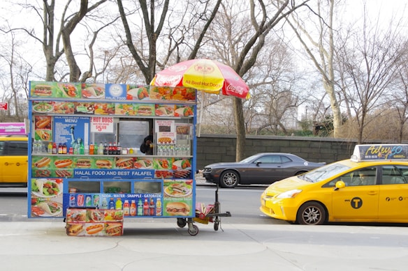 A street-side halal food cart offering a variety of items like hot dogs and falafel. The cart is adorned with colorful food images and a bright umbrella. It is parked on a city street next to a yellow taxi and a gray car, with leafless trees and a stone wall in the background.