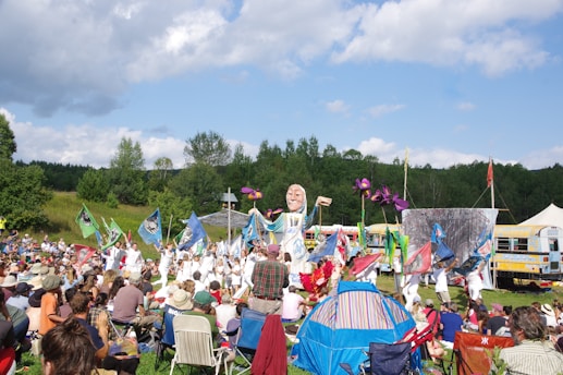 A large outdoor gathering with people sitting and watching a performance. There are colorful flags and a giant puppet among the participants. The setting is a grassy field surrounded by trees, with school buses and tents in the background. The sky is partly cloudy with plenty of sunshine.
