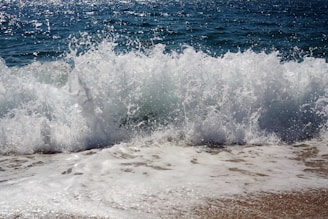 Waves crashing onto a sandy shore with sea spray and foam. The ocean water appears deep blue, and the motion of the waves is prominently captured.