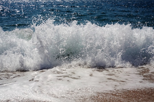 Waves crashing onto a sandy shore with sea spray and foam. The ocean water appears deep blue, and the motion of the waves is prominently captured.
